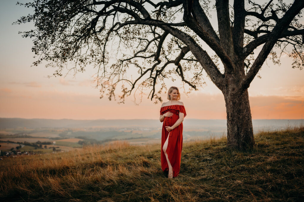 Das Bild zeigt eine schwangere Frau in einem roten langen Kleid. Sie umfasst mit beiden Händen liebevoll ihren Babybauch.