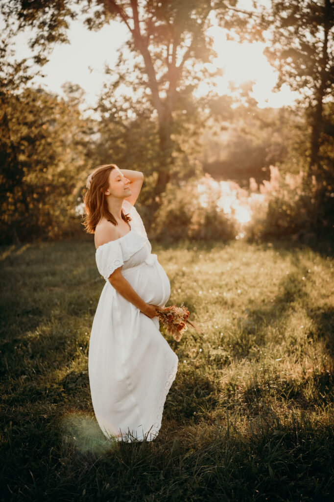 Das Bild zeigt eine wunderschöne schwangere Frau in einem weißen Kleid auf einer Wiese vor der untergehenden Sonne.
