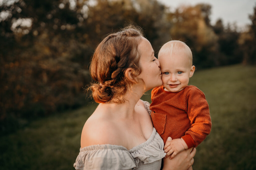 Das Bild zeigt eine Mama, die ihr Kind auf dem Arm hat und ihm einen Kuss auf die Schläfe gibt.