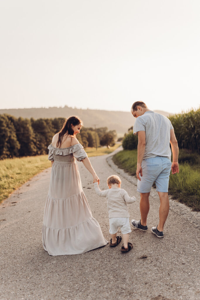 Das Bild zeigt eine Mama, in einem bodenlangen Kleid und einen Papa, die ihr Kleinkind an den Händen zwischen sich halten. Die Familie läuft vom Betrachter weg.