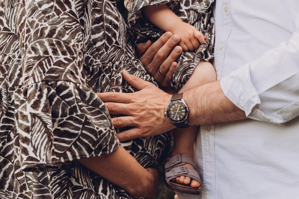 Das Bild zeigt den schwangeren Bauch einer Mama und die Hände des Papas, der ihn berührt und der gleichzeitig ein Kind im Arm trägt.