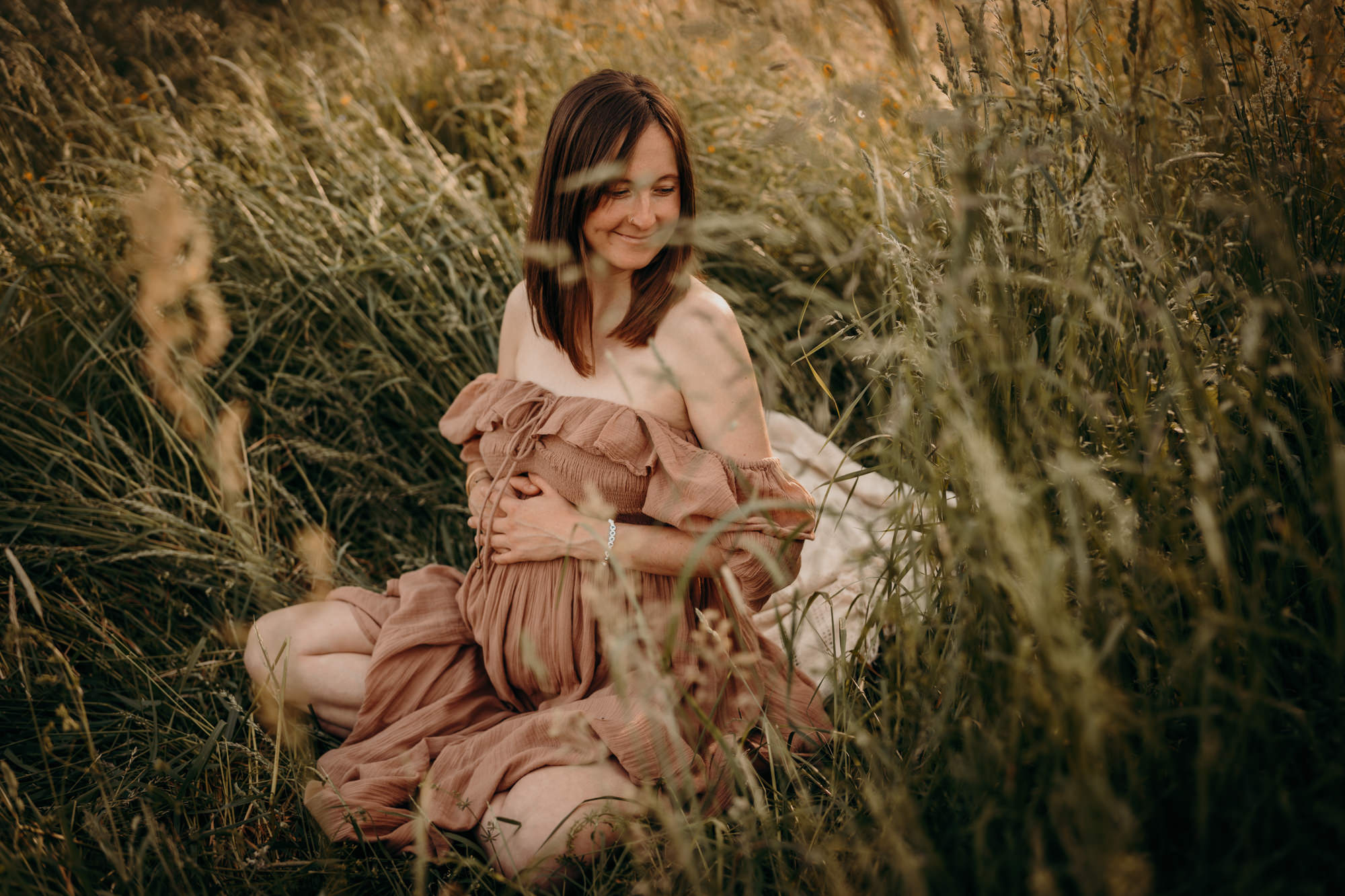 Das Bild zeigt eine schwangere Frau in einem Kleid im hohen Gras sitzend.