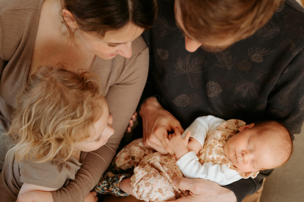 Das Bild zeigt ein Neugeborenes im Arm der Eltern, die es bewundernd gemeinsam mit dem älteren Geschwisterchen anschauen.